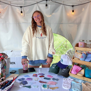Person standing behind a table with various items at an outdoor event