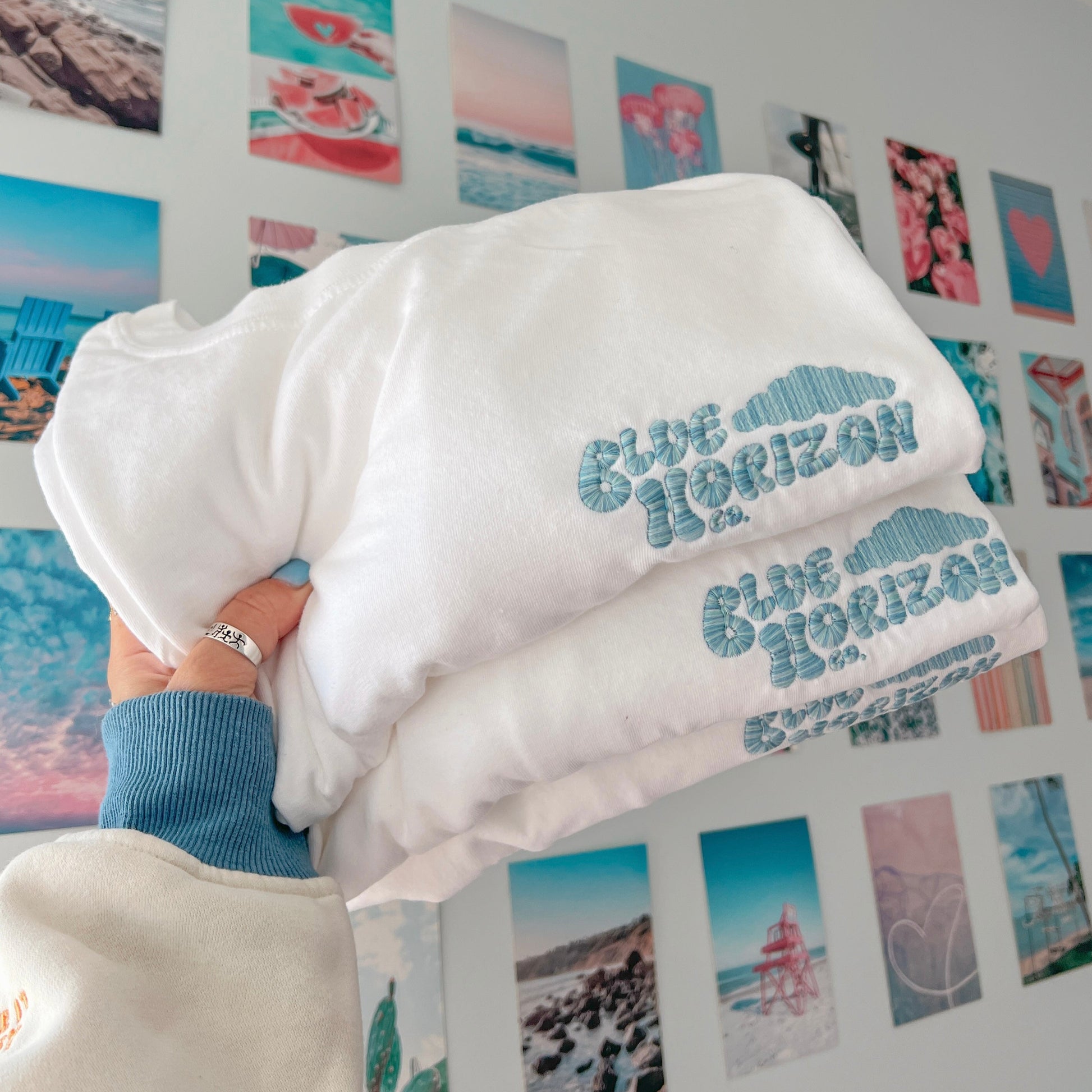 White hoodie with 'Blue Horizon' text held in front of a wall with beach-themed prints.