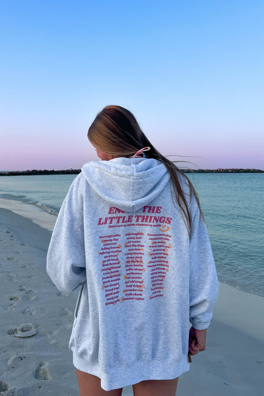 A person standing on a beach at sunset wearing a gray zip-up hoodie with text on the back.