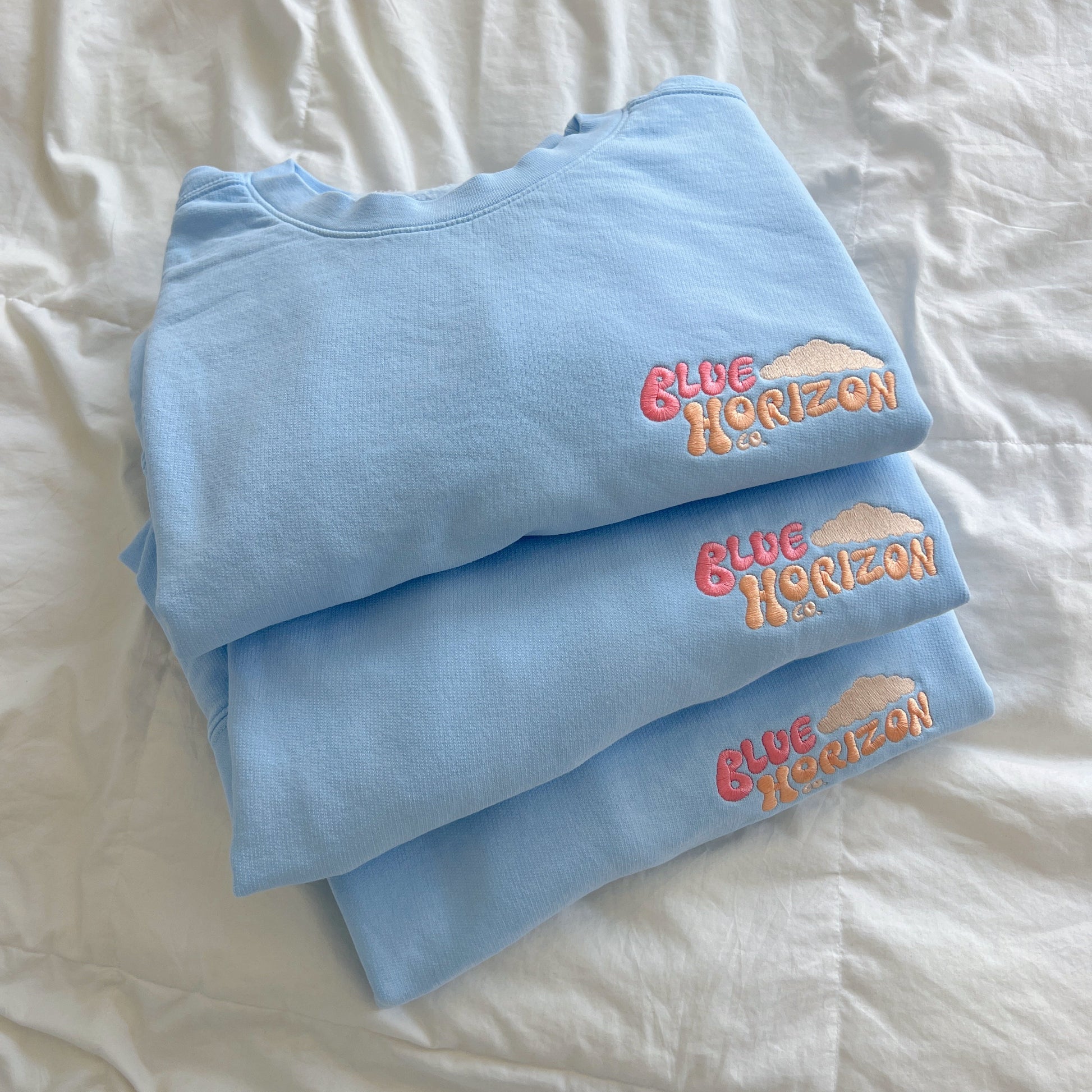 Folded light blue sweatshirts with 'Blue Horizon' text on a white bedspread